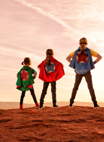 Three kids in superhero capes standing on a hilltop at sunset