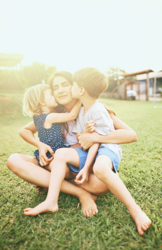 A smiling mom sitting in the grass while her two kids kiss her cheeks